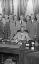 President Truman is seated behind a desk signing a document with men standing behind him.