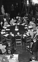 Delegates from the United States, Great Britain, and the Soviet Union sit around a conference table, surrounded by aides and photographers. 